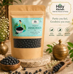 A stand-up kraft pouch of Holy Kitchen Premium Hingwati herbal capsules displayed on a rustic stone pedestal with bowls of blueberries and decorative props in a warm, styled setting.